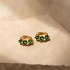 Triple Emerald Huggies: gold hoop earrings with three green gemstones each, shown on a beige textured surface in soft light—perfect personalized jewelry for any occasion.