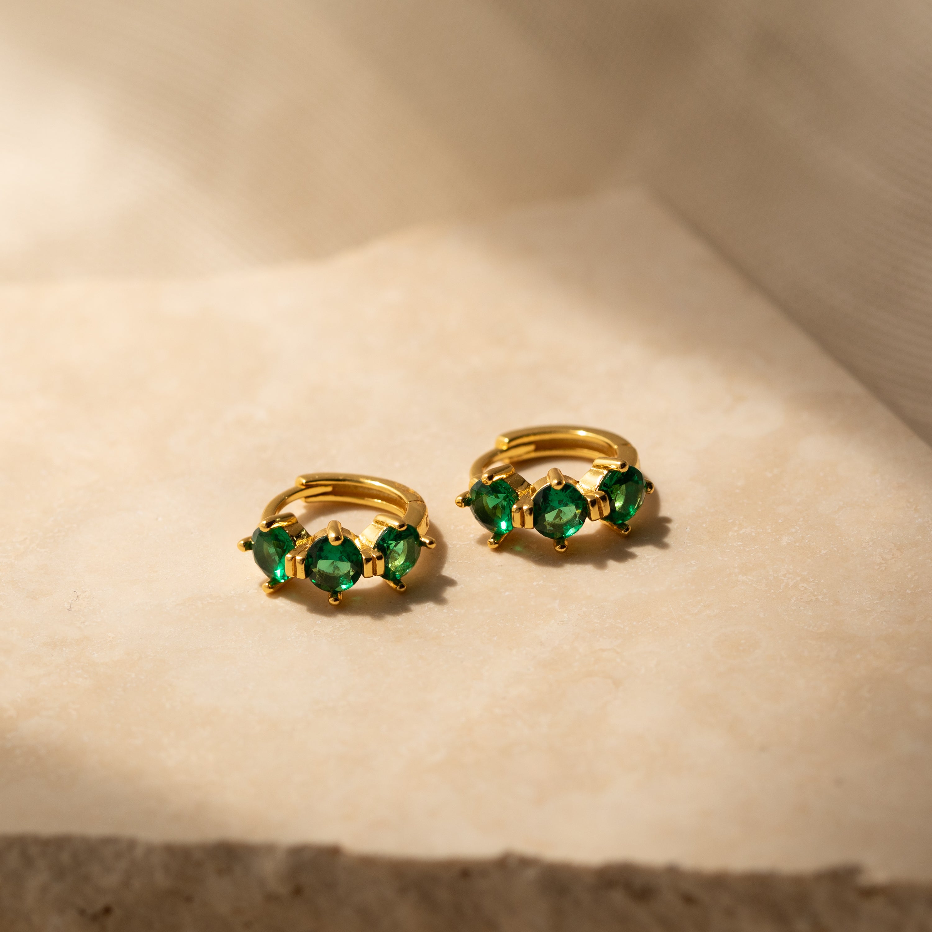 Triple Emerald Huggies: gold hoop earrings with three green gemstones each, shown on a beige textured surface in soft light—perfect personalized jewelry for any occasion.