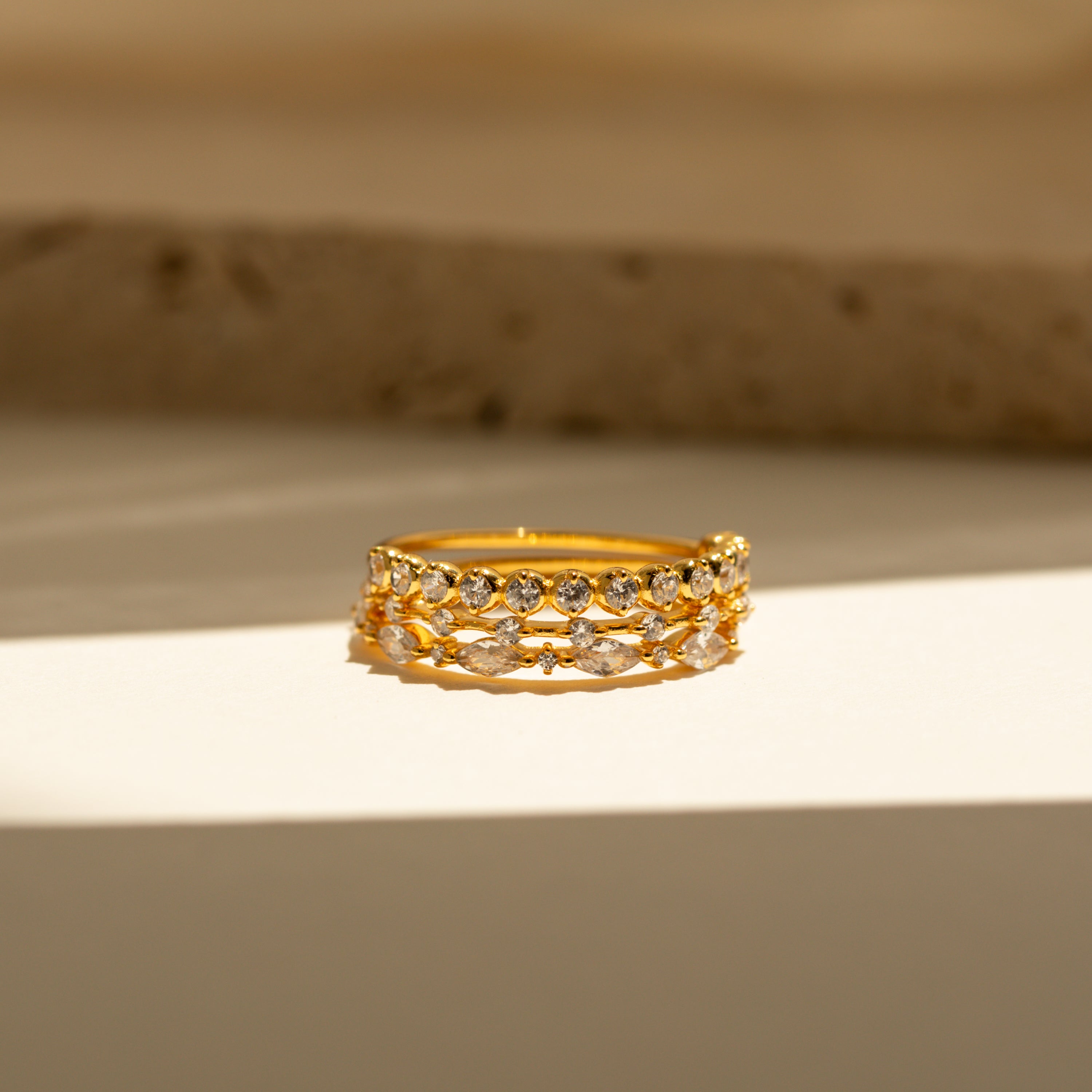 The Crystal Stacking Ring Set in 18K Gold features two rows of sparkling crystal gemstones, elegantly displayed on a sunlit surface with a neutral background—a stunning jewelry piece.