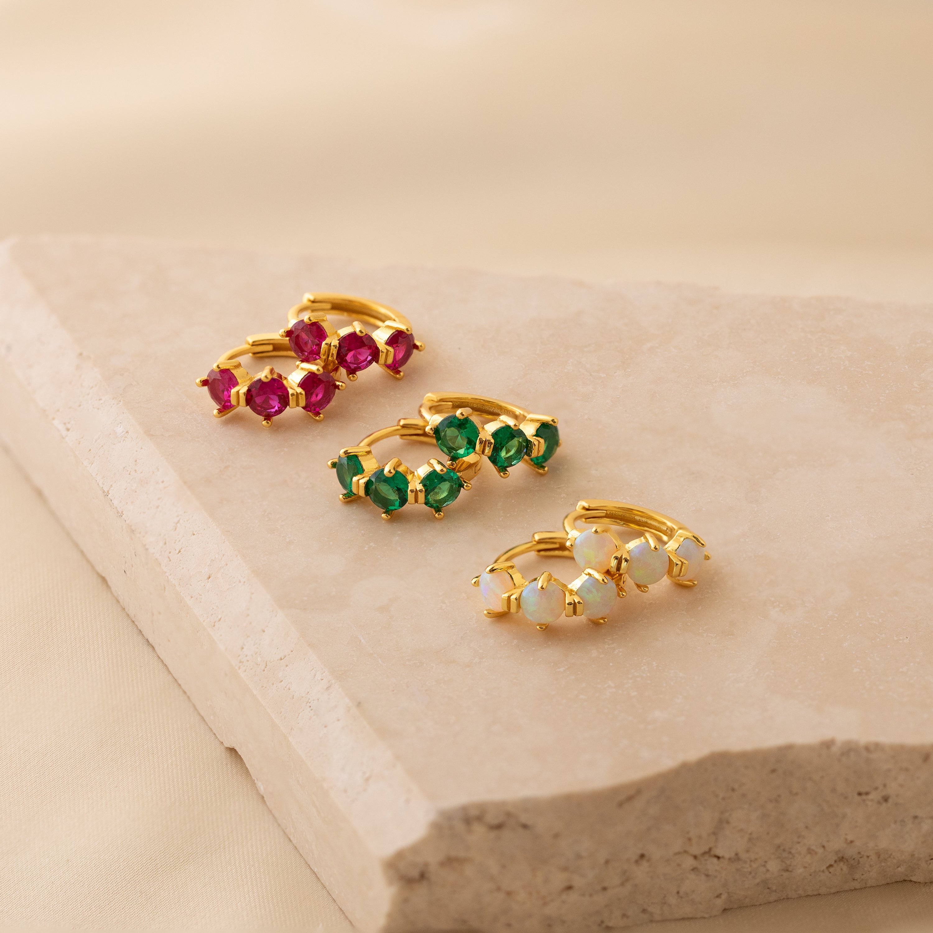 Three pairs of minimalist hoops—Triple Emerald Huggies—featuring gold and green stones rest on a beige stone slab, perfect for lovers of personalized jewelry.