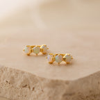 Triple Opal Birthstone Huggies feature three round opal stones in a minimalist gold design, beautifully displayed on a textured beige stone surface.