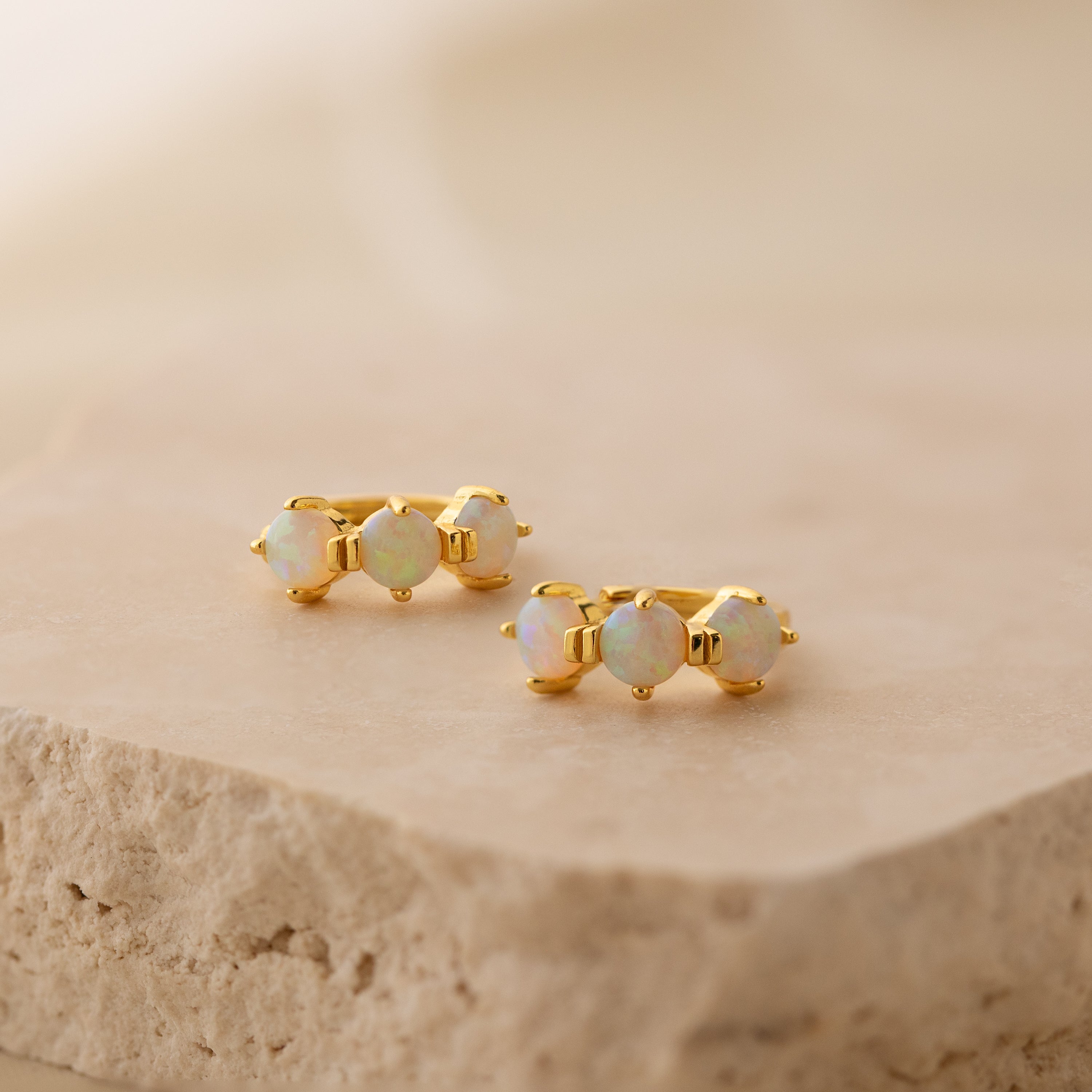 Triple Opal Birthstone Huggies feature three round opal stones in a minimalist gold design, beautifully displayed on a textured beige stone surface.