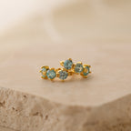 The Triple Aquamarine Huggies feature clusters of light blue gemstones on gold hoops, displayed atop a beige stone—perfect minimalist earrings customizable with birthstones.