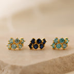 Three pairs of gold earrings—minimalist hoops and Triple Blue Zircon Huggies—featuring blue and green gemstones, displayed on a beige stone surface. These customizable gemstone earrings add a refined touch to any style.