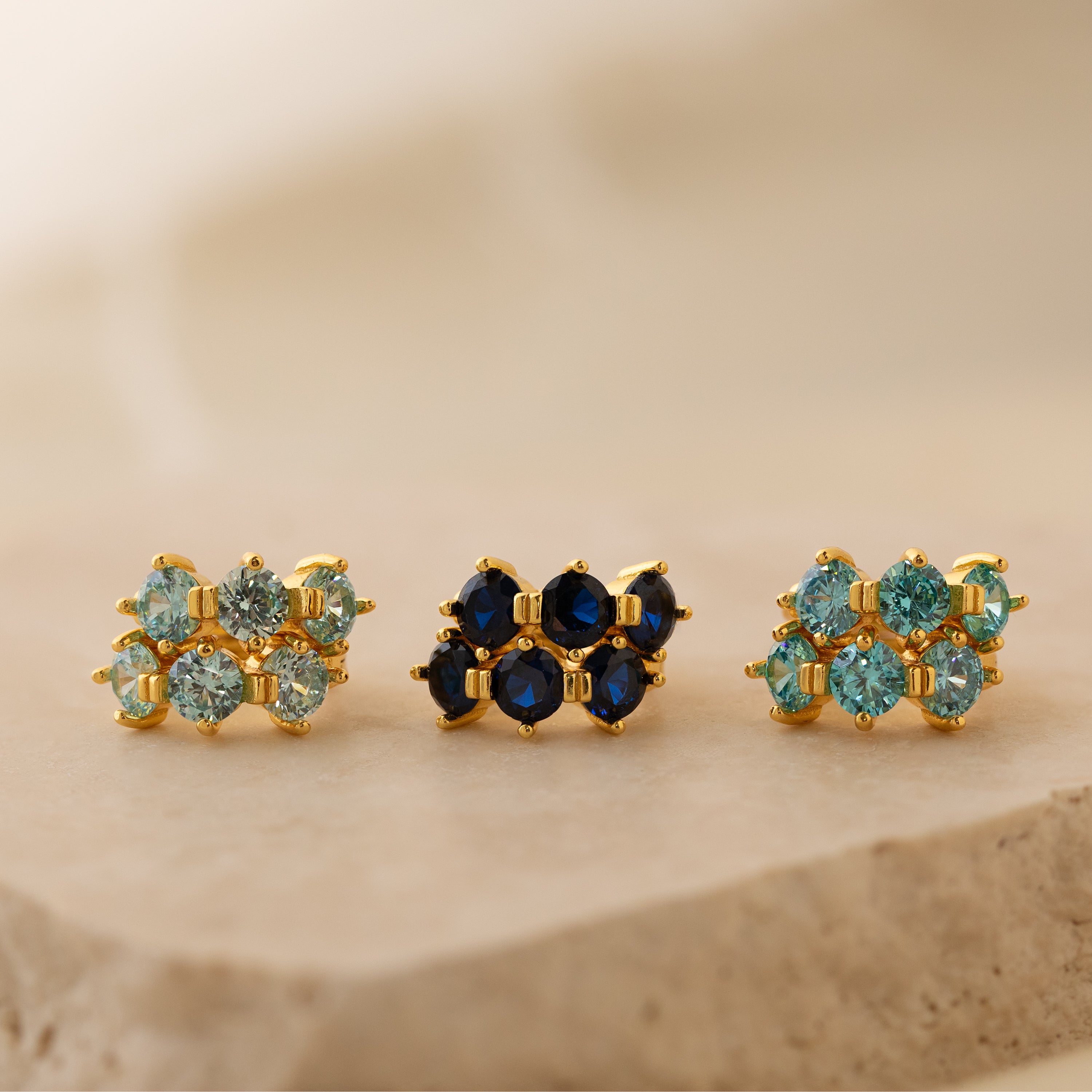 Three pairs of gold earrings—minimalist hoops and Triple Blue Zircon Huggies—featuring blue and green gemstones, displayed on a beige stone surface. These customizable gemstone earrings add a refined touch to any style.