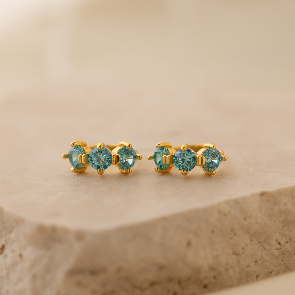 Triple Blue Zircon Huggies featuring three round blue zircon stones on a beige stone surface—perfect customizable gemstone earrings for minimalist hoop lovers.