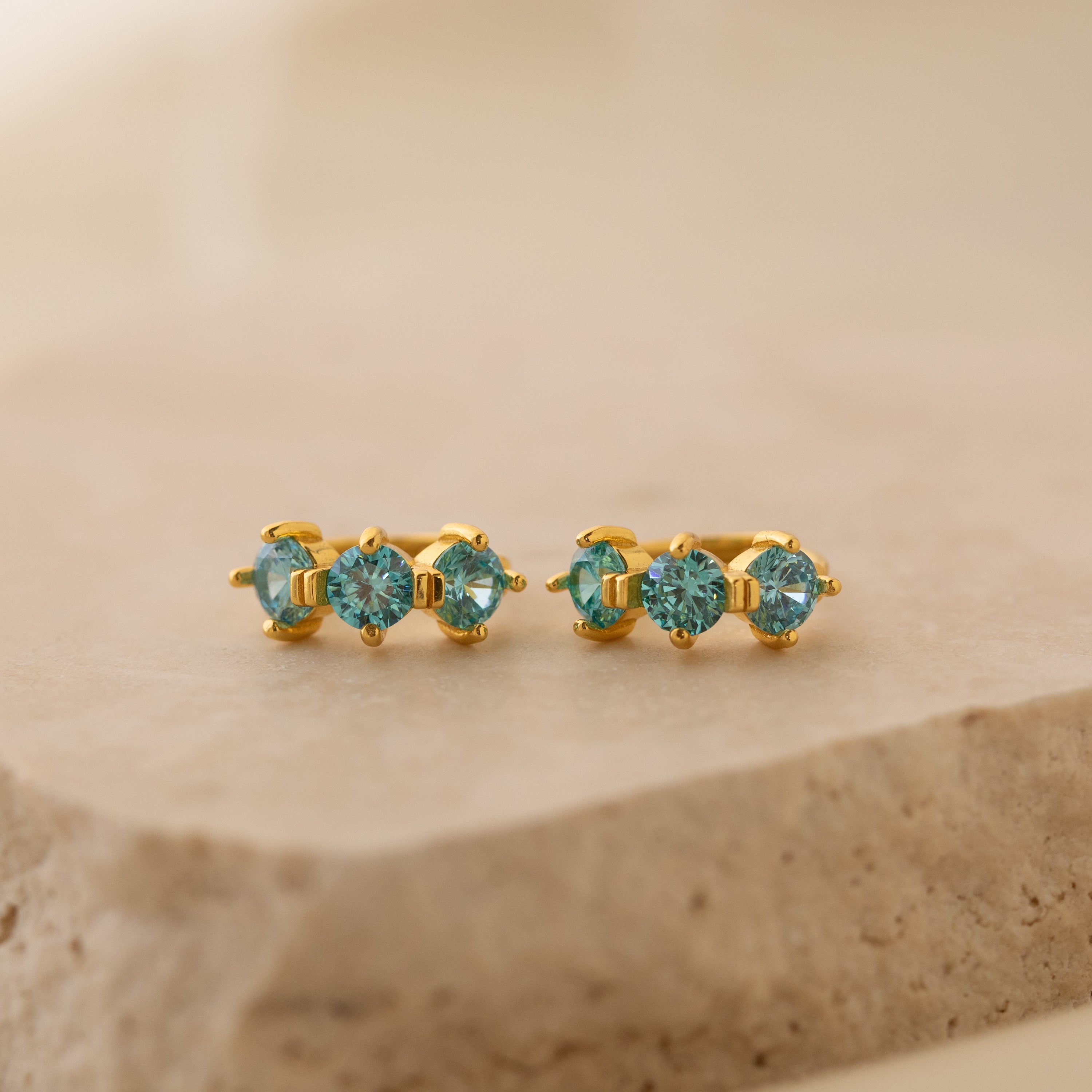 Triple Blue Zircon Huggies featuring three round blue zircon stones on a beige stone surface—perfect customizable gemstone earrings for minimalist hoop lovers.