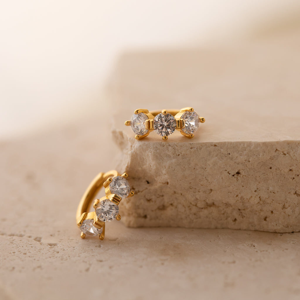 Triple Diamond Huggies gold earrings feature three sparkling diamonds each, displayed on a light beige stone surface—an ideal pick for minimalist jewelry lovers.