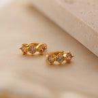 Triple Topaz Huggies feature three sparkling round topaz gemstones on each gold earring, displayed on soft beige fabric—minimalist and customizable hoops for a personalized touch.