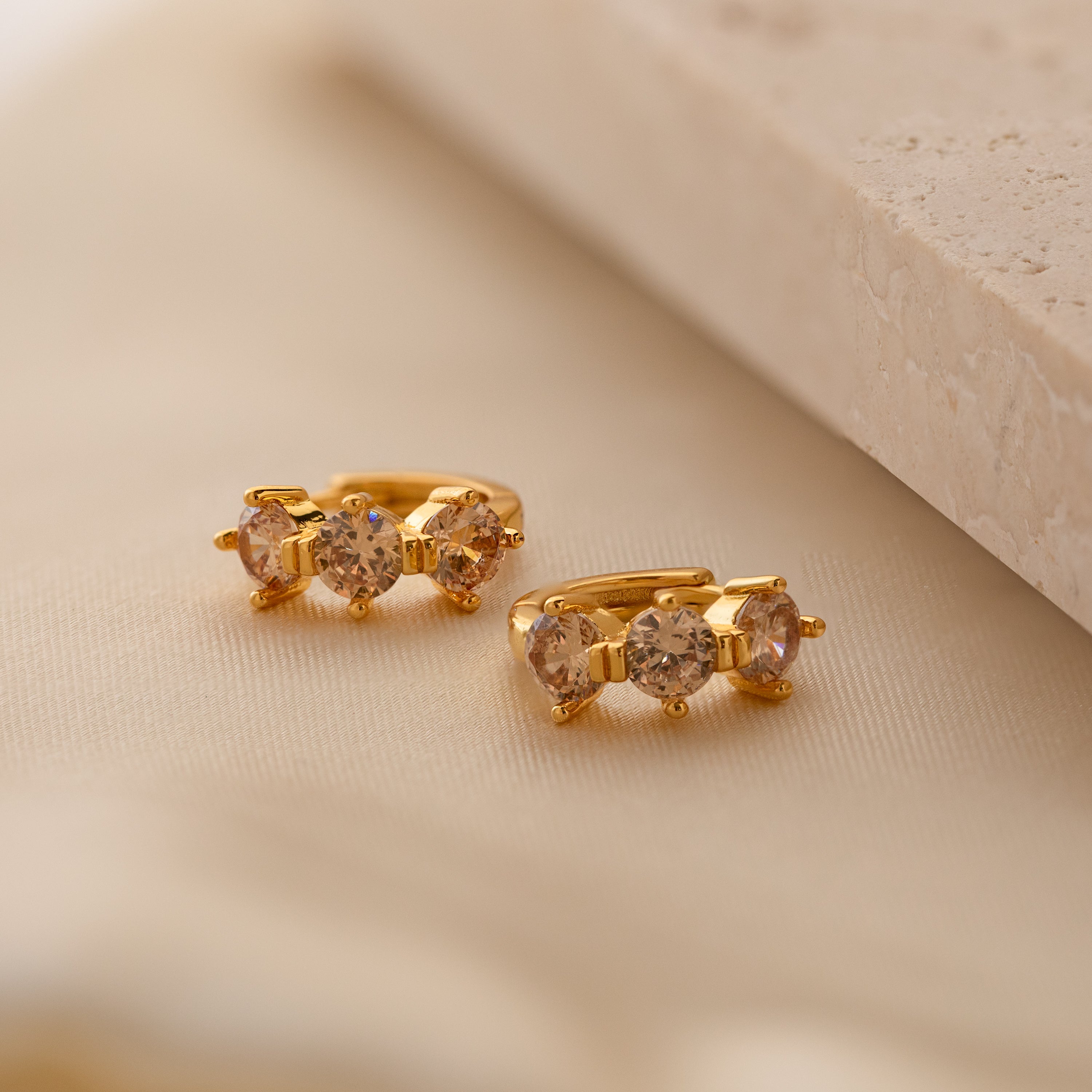 Triple Topaz Huggies feature three sparkling round topaz gemstones on each gold earring, displayed on soft beige fabric—minimalist and customizable hoops for a personalized touch.