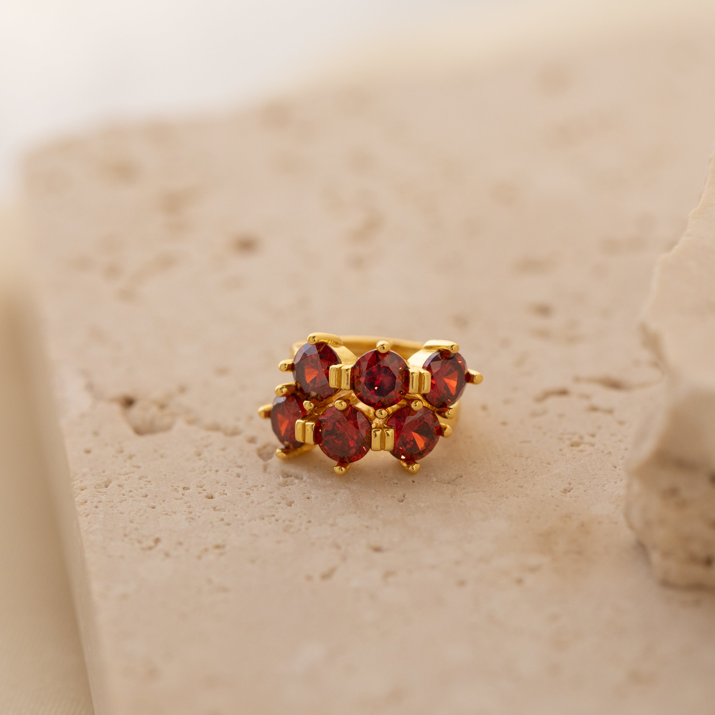 The Triple Garnet Huggies, gold rings featuring six red garnets, are displayed on a textured beige stone surface, blending minimalist hoop elegance with the personalized allure of customizable birthstone earrings.