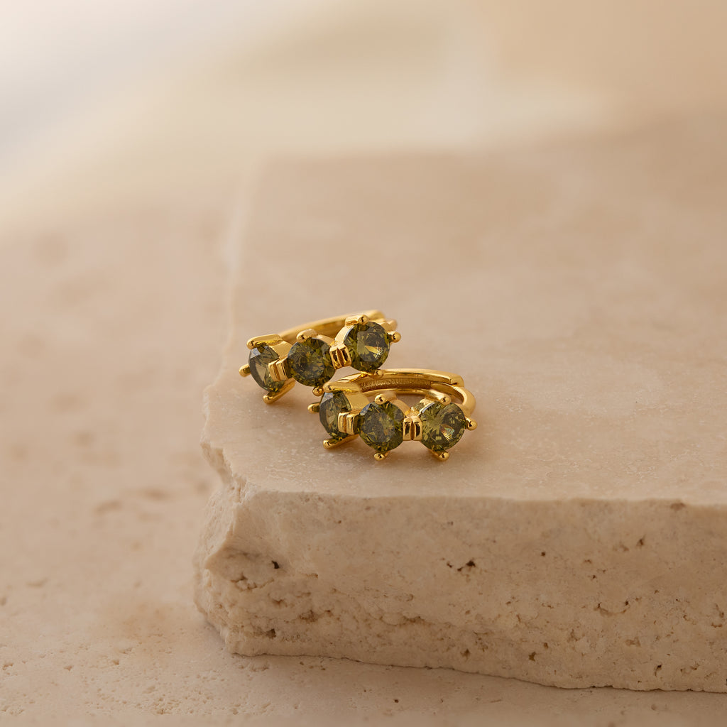 Triple Peridot Huggies: Minimalist gold hoops accented with green peridot stones, elegantly displayed on a textured beige stone surface.