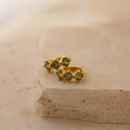 Triple Peridot Huggies: Minimalist gold hoops accented with green peridot stones, elegantly displayed on a textured beige stone surface.