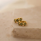 Triple Peridot Huggies: Minimalist gold hoops accented with green peridot stones, elegantly displayed on a textured beige stone surface.