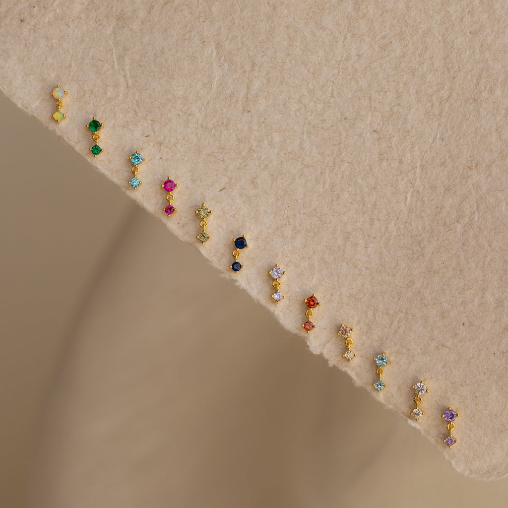 Birthstone Drop Studs, available in vibrant pairs, are displayed on a textured beige fabric—perfect for unique birth month jewelry gifts.