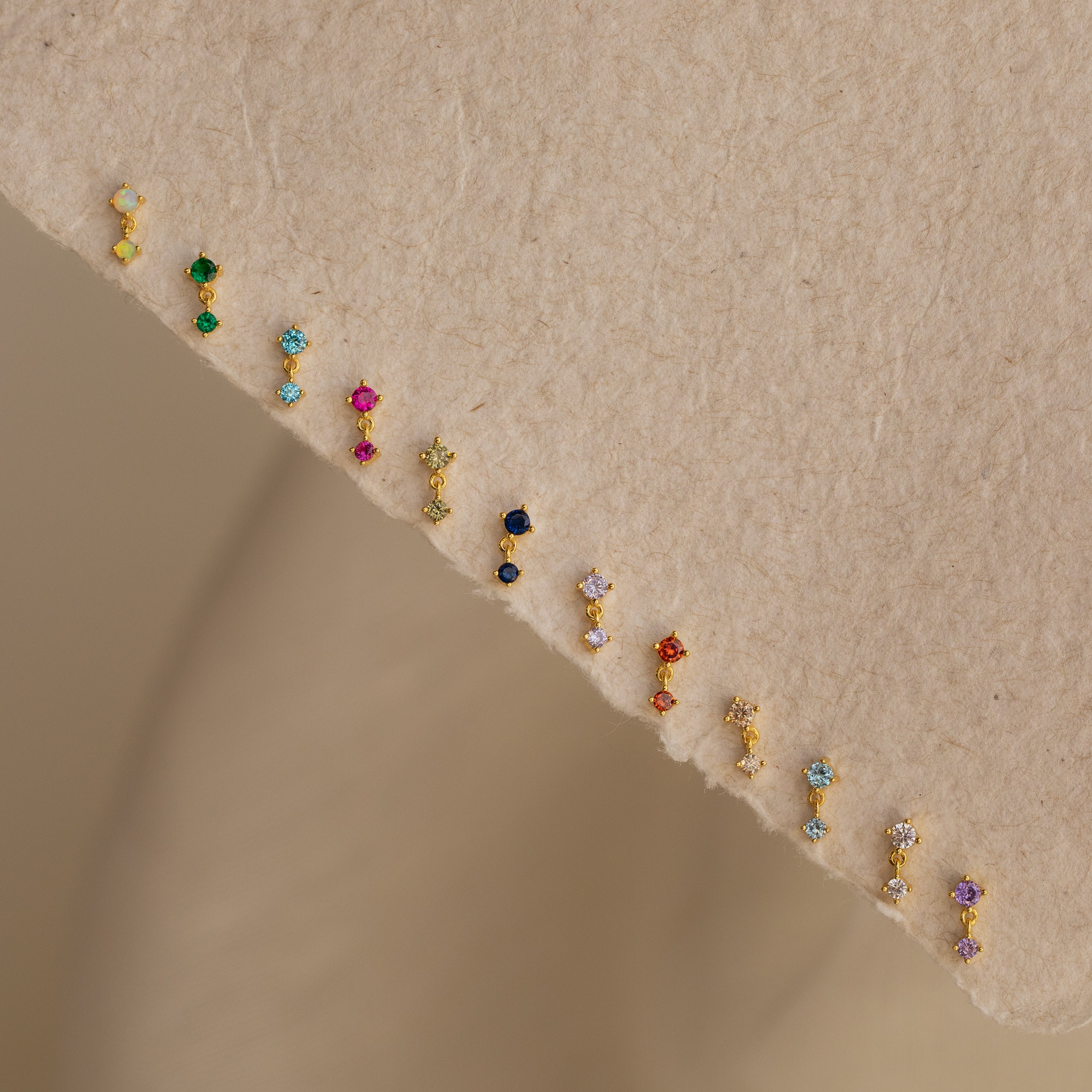 Birthstone Drop Studs, available in vibrant pairs, are displayed on a textured beige fabric—perfect for unique birth month jewelry gifts.