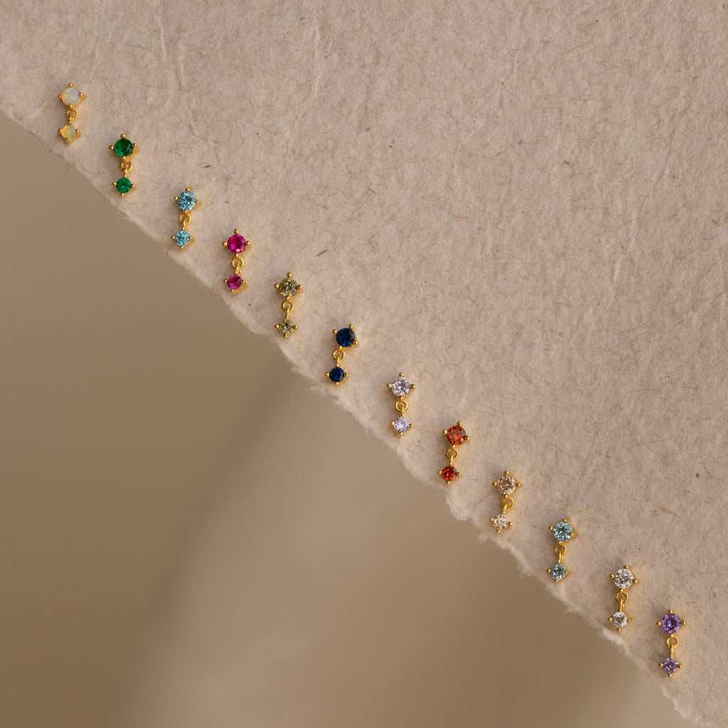 A row of sparkling Diamond Drop Studs is arranged on a textured beige surface, making each accessory a dazzling highlight for any jewelry collection.