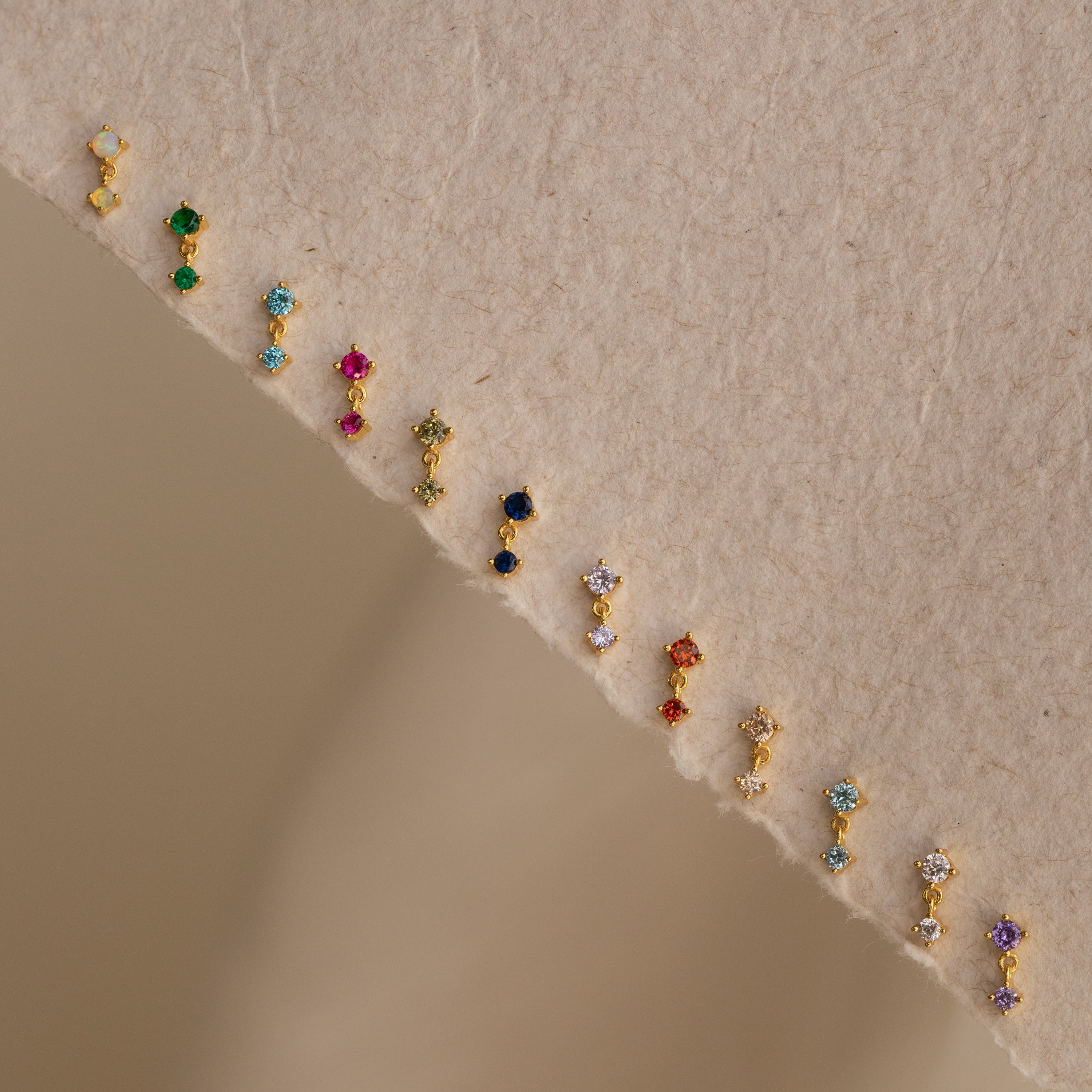 A row of sparkling Diamond Drop Studs is arranged on a textured beige surface, making each accessory a dazzling highlight for any jewelry collection.