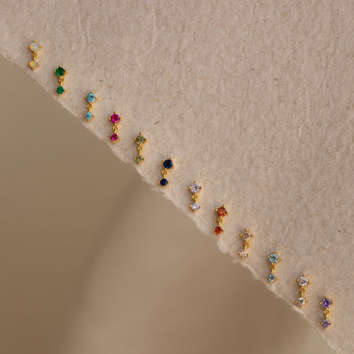 The Birthstone Drop Studs are displayed in a row on a textured beige surface, highlighting vibrant gemstone earrings that add a perfect touch to any occasion.