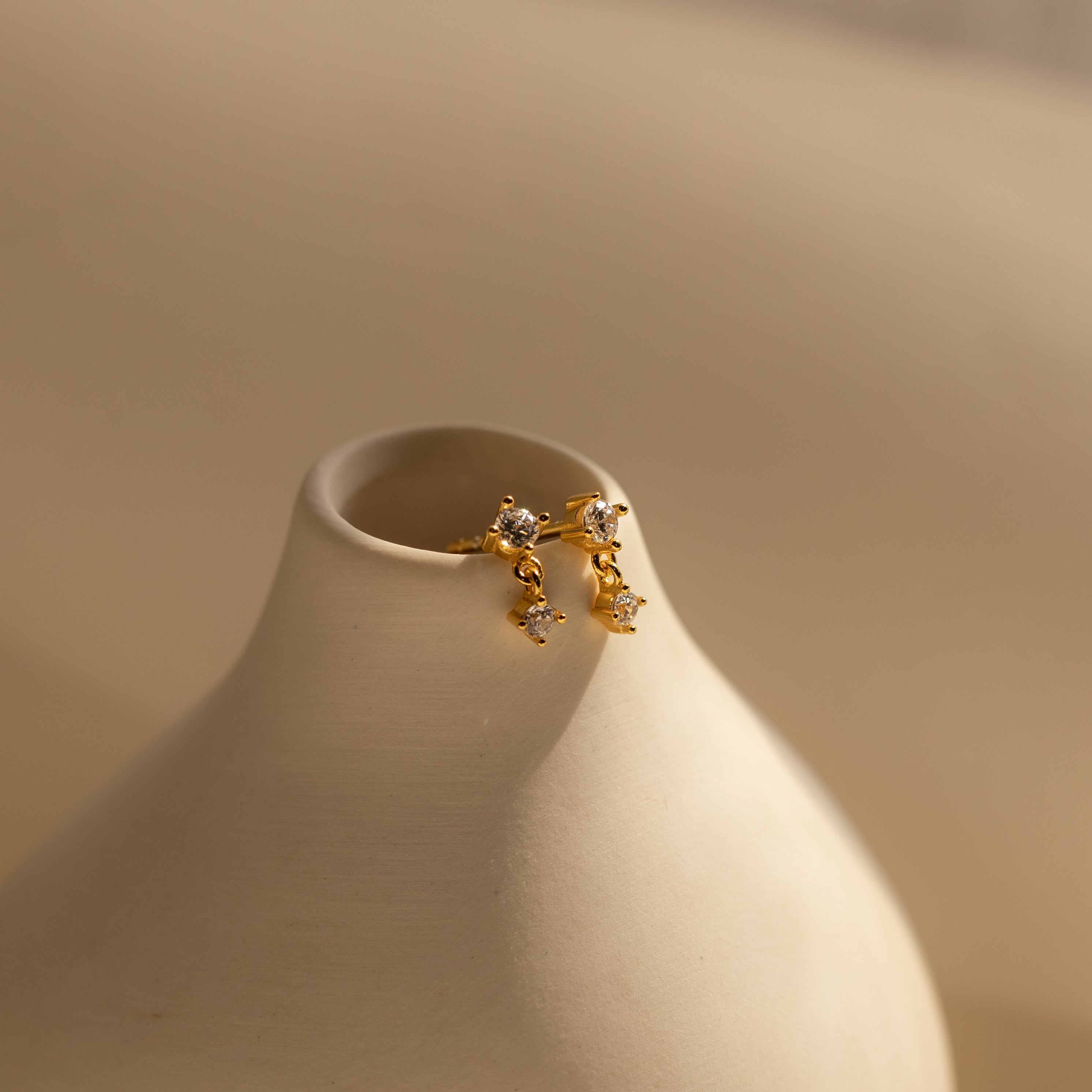 Topaz Drop Studs birthstone earrings with small crystals in gold and silver, a chic accessory, shown on a smooth cream ceramic vase.