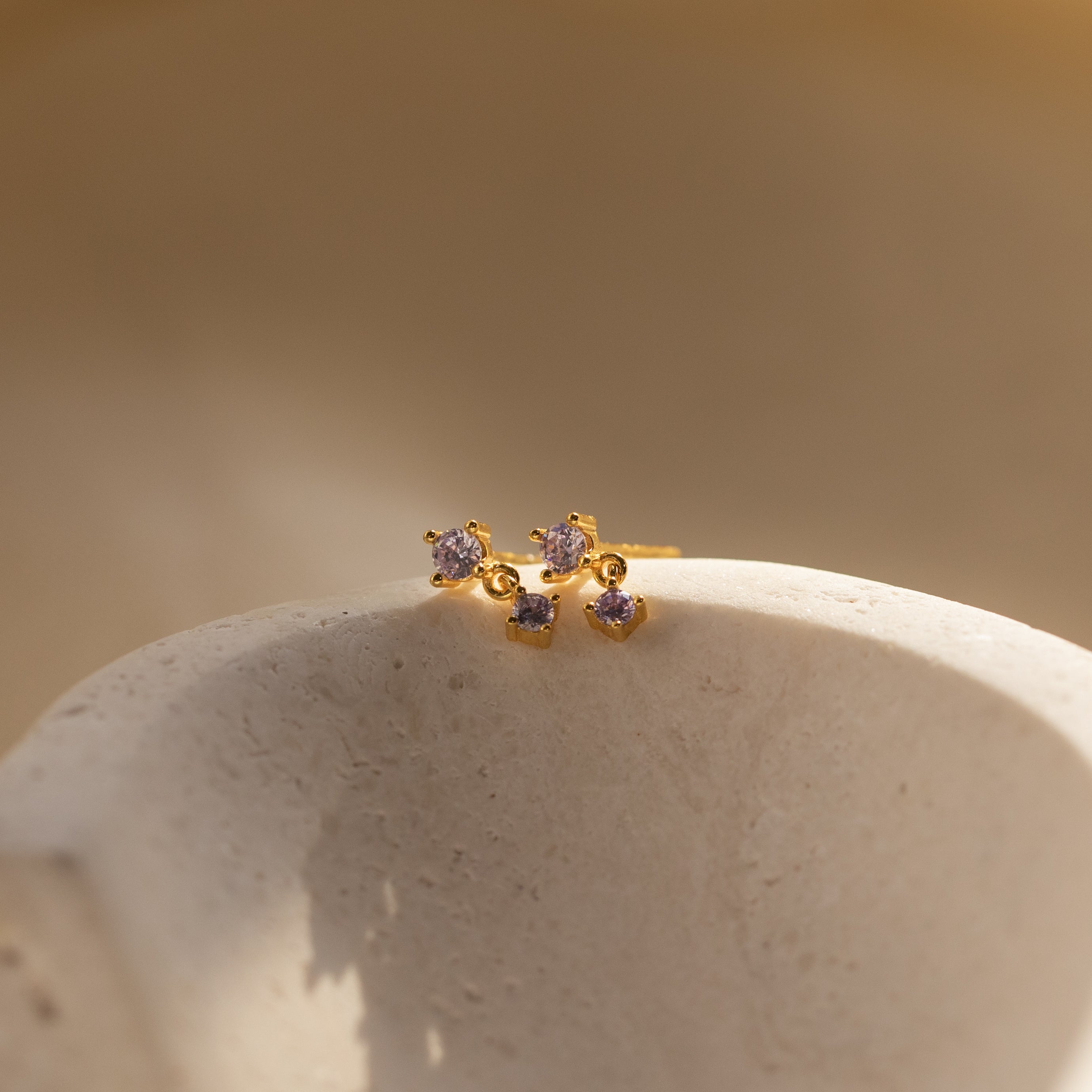 The Alexandrite Drop Studs, featuring two gold gemstone studs with small purple stones, are displayed on a textured, off-white curved surface.