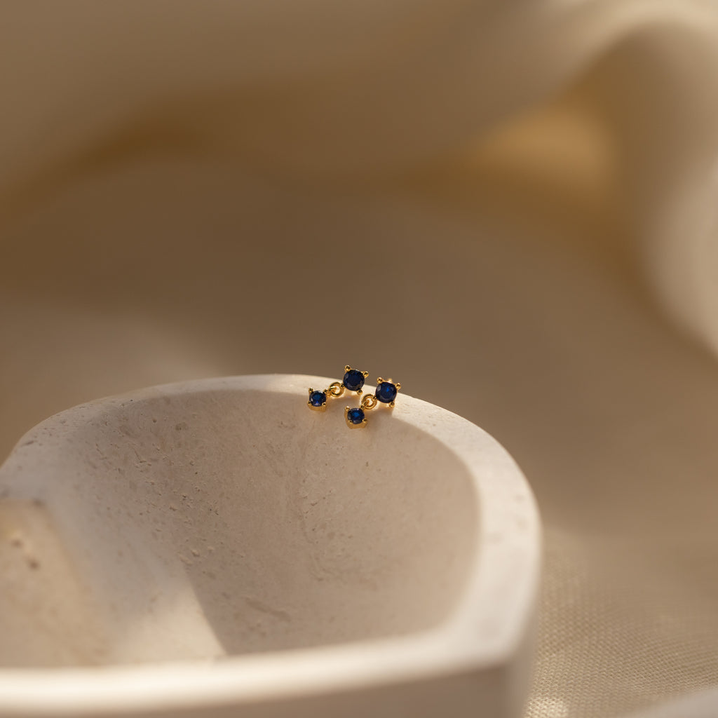 Sapphire Drop Studs with small blue gemstones rest on a smooth, white stone surface against a soft beige background—an elegant gold birthstone earring perfect for any occasion.