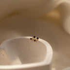 Sapphire Drop Studs with small blue gemstones rest on a smooth, white stone surface against a soft beige background—an elegant gold birthstone earring perfect for any occasion.