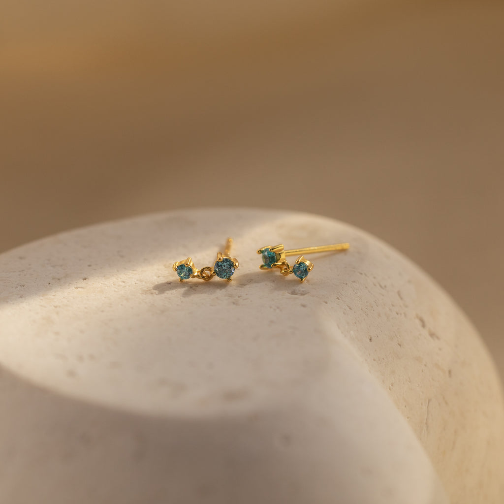 Blue Zircon Drop Studs with small blue stones rest on a smooth, light-colored stone surface in soft lighting.