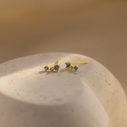 Blue Zircon Drop Studs with small blue stones rest on a smooth, light-colored stone surface in soft lighting.