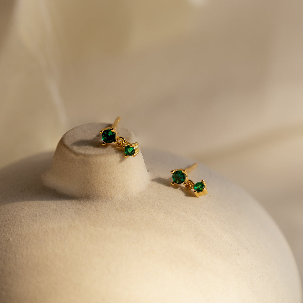 Birthstone Drop Studs featuring green gemstones set in gold, displayed on a beige, rounded surface with a soft background.