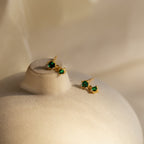 Birthstone Drop Studs featuring green gemstones set in gold, displayed on a beige, rounded surface with a soft background.