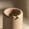 Emerald Birthstone Drop Studs, featuring dainty green gemstones, are showcased on a smooth beige cylindrical stand in soft light—perfect as elegant birthstone drop earrings.