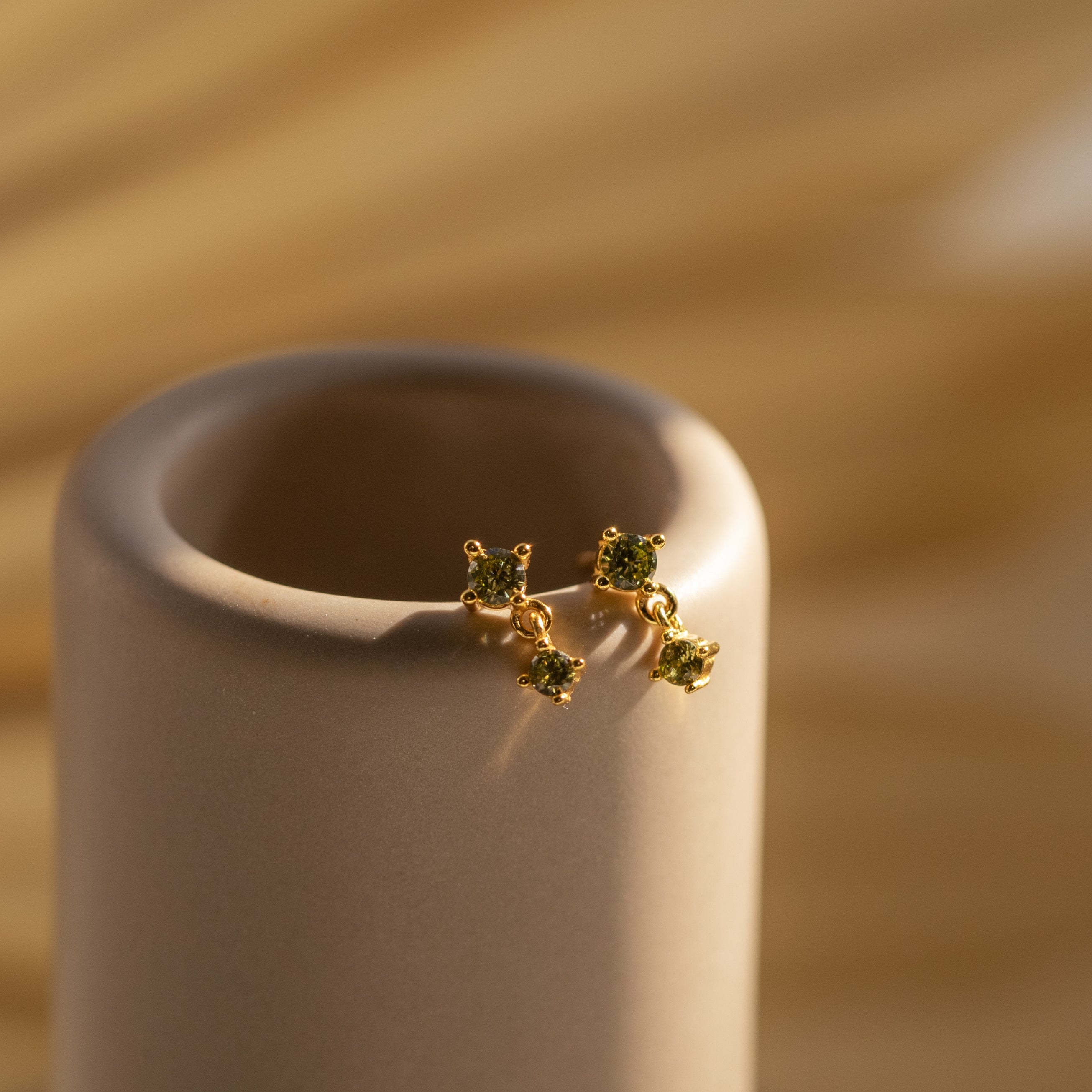 Two Peridot Drop Studs with petite gems rest on a smooth, beige cylindrical surface in soft light.