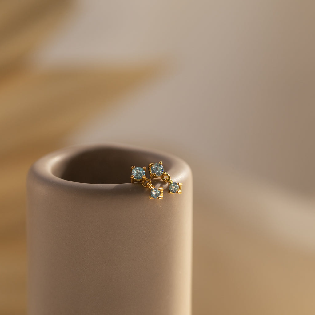 Aquamarine Drop Studs with petite blue gems rest on a smooth, beige cylindrical stand.