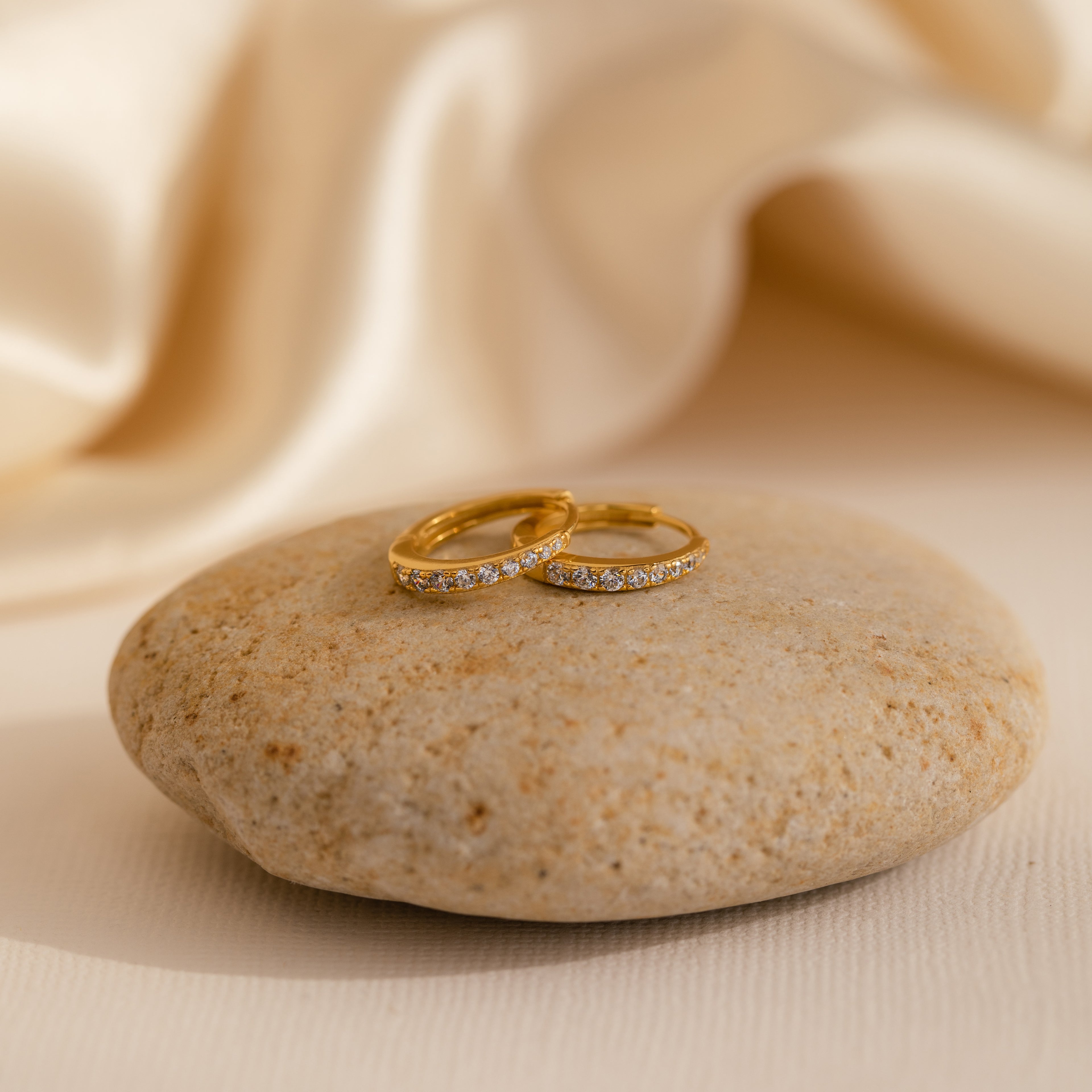The Vera Pave Diamond Huggies and two gold rings with small diamonds are elegantly displayed on a smooth stone, set against a soft cream fabric background.