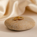The Vera Pave Garnet Huggies, two gold rings set with vivid red gemstones, rest on a smooth beige stone against a soft cream fabric background, adding an elegant touch reminiscent of fine gemstone earrings.