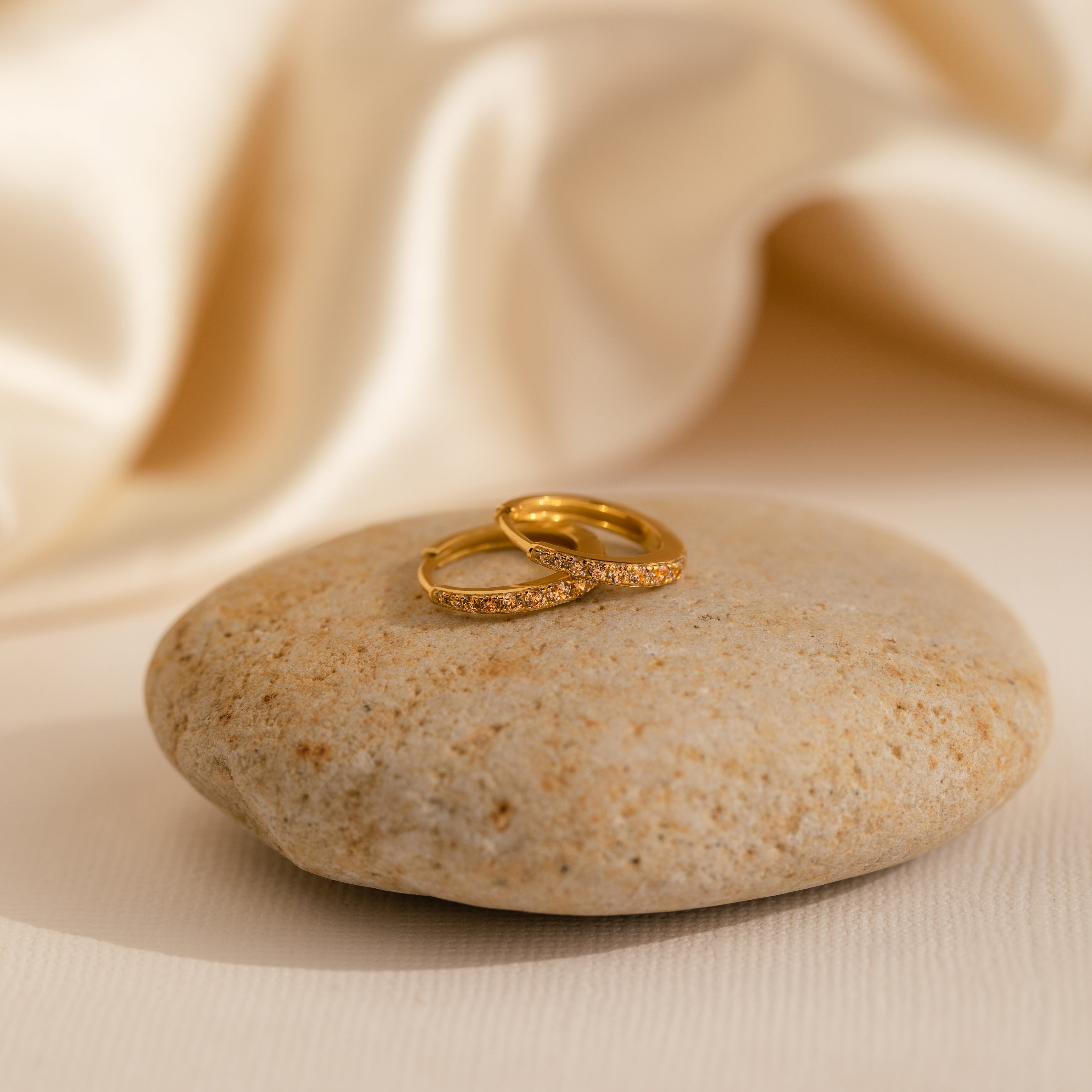 Two gold rings with small diamonds rest on a smooth stone, while the Vera Pave Topaz Huggies add a vibrant touch against the soft cream fabric background.