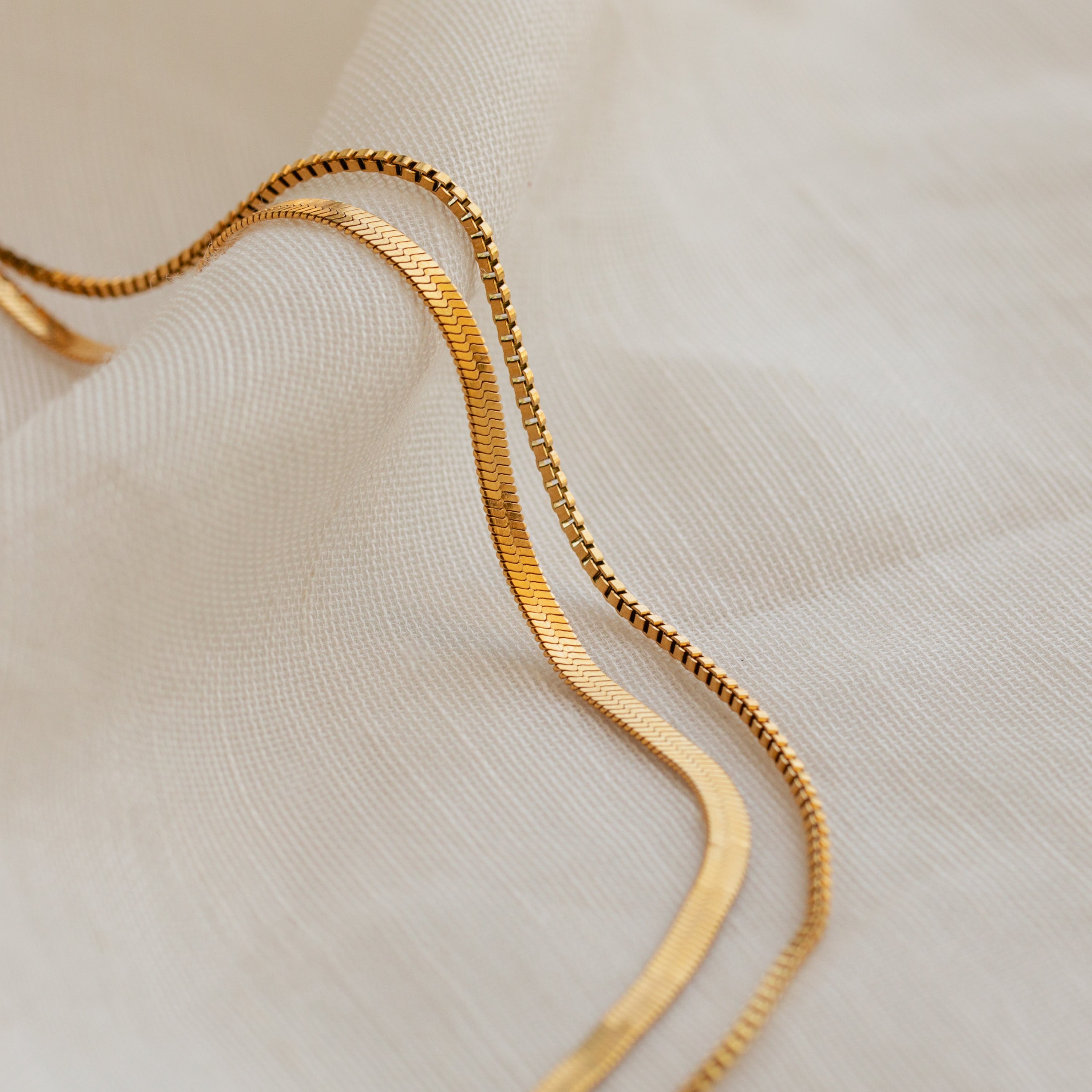 The Herringbone Duo Chain Necklace in 18K Gold rests on soft, white textured fabric with gentle, wavy folds.