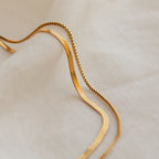 The Herringbone Duo Chain Necklace rests in soft curves on a textured fabric background, highlighting its elegant, minimalist design.
