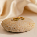 The Vera Pave Peridot Huggies, gold rings adorned with small peridot gemstones, rest on a smooth beige stone and are paired with soft cream fabric for an elegant, timeless look.