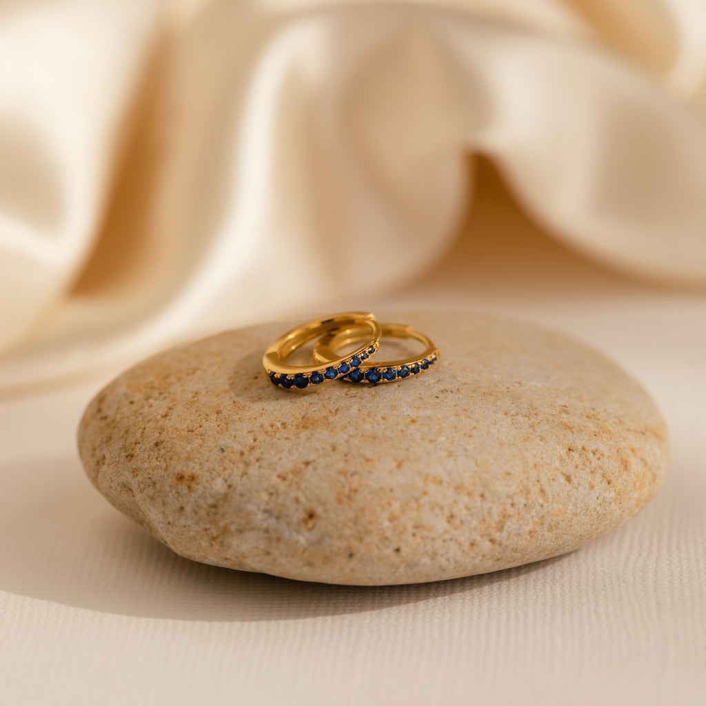 The Vera Pave Sapphire Huggies, gold rings set with blue stones, are displayed on a smooth beige rock against a soft, cream-colored fabric background.