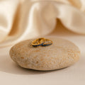 The Vera Pave Sapphire Huggies, gold rings set with blue stones, are displayed on a smooth beige rock against a soft, cream-colored fabric background.