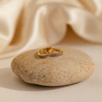The Vera Pave Amethyst Huggies, two gold rings adorned with small purple amethyst gems, rest on a smooth stone against a backdrop of cream-colored fabric.