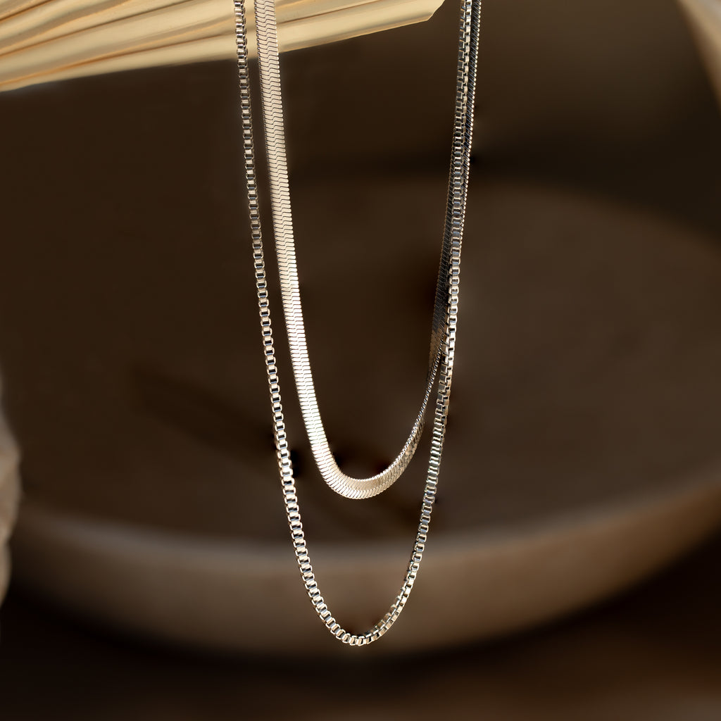 The Herringbone Duo Chain Necklace, featuring two delicate silver chains, drapes elegantly over an open book set against a soft, neutral background.