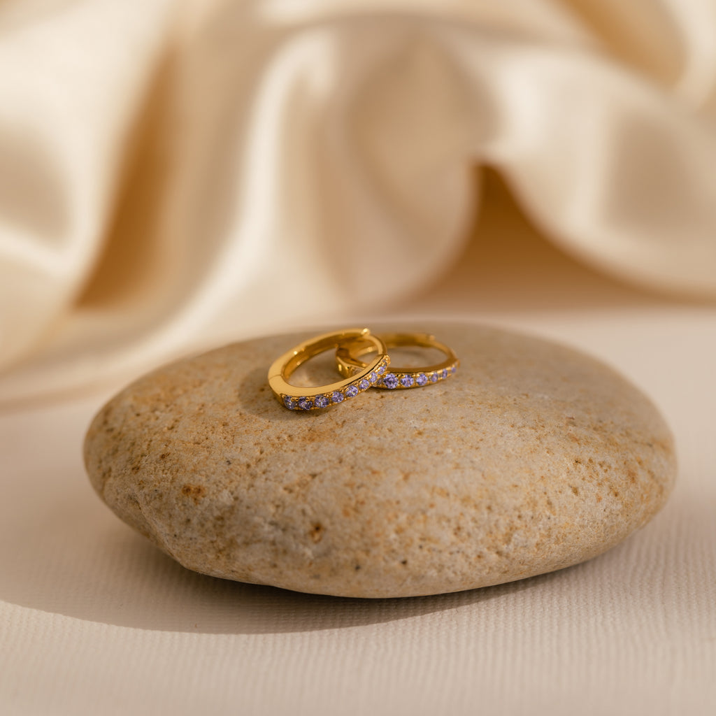 The Vera Pave Alexandrite Huggies, featuring sparkling stones and a gold finish, are displayed on a smooth beige stone against a soft fabric background.