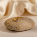 The Vera Pave Alexandrite Huggies, featuring sparkling stones and a gold finish, are displayed on a smooth beige stone against a soft fabric background.