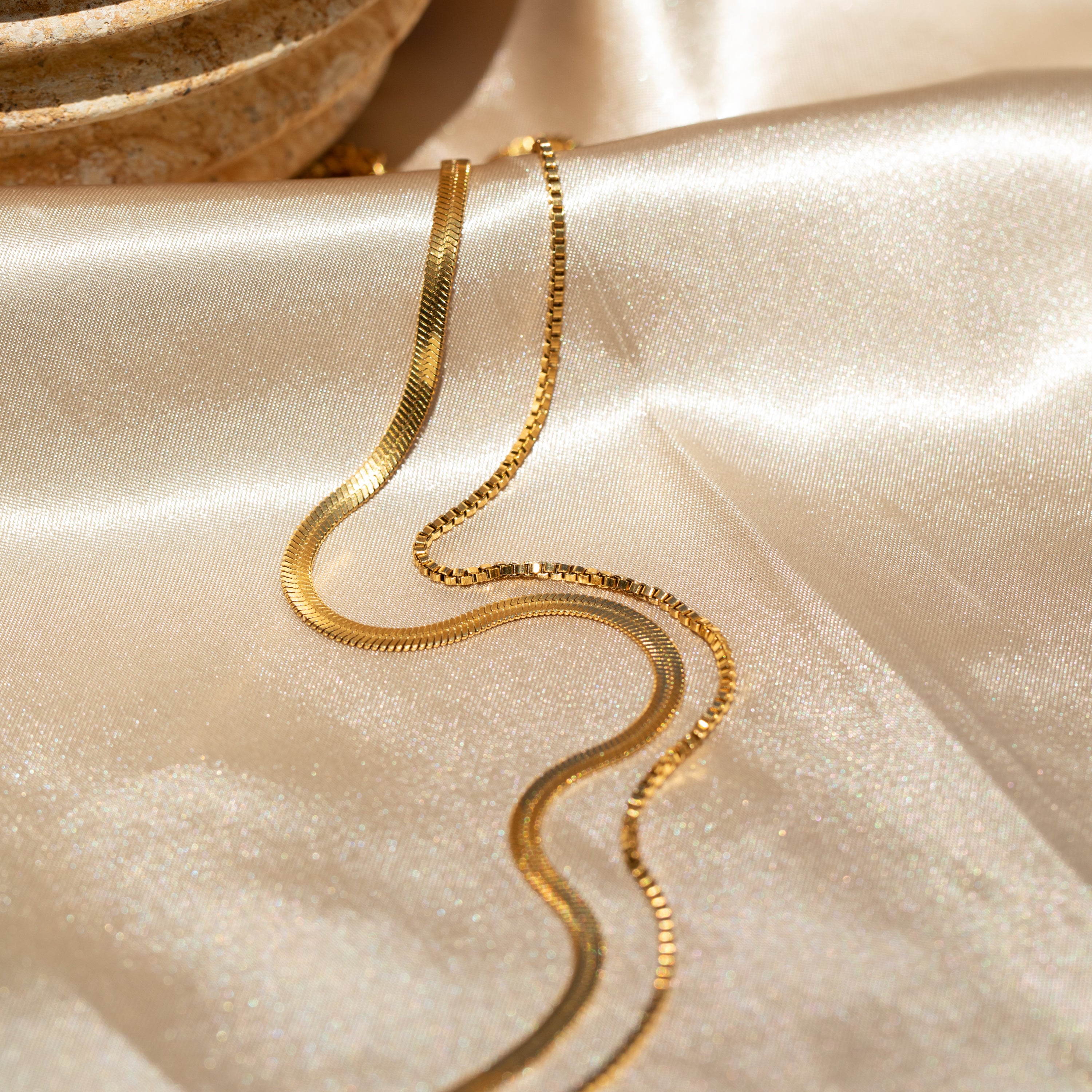 The Herringbone Duo Chain Necklace curves elegantly on shiny beige satin, with a stone bowl in the corner.