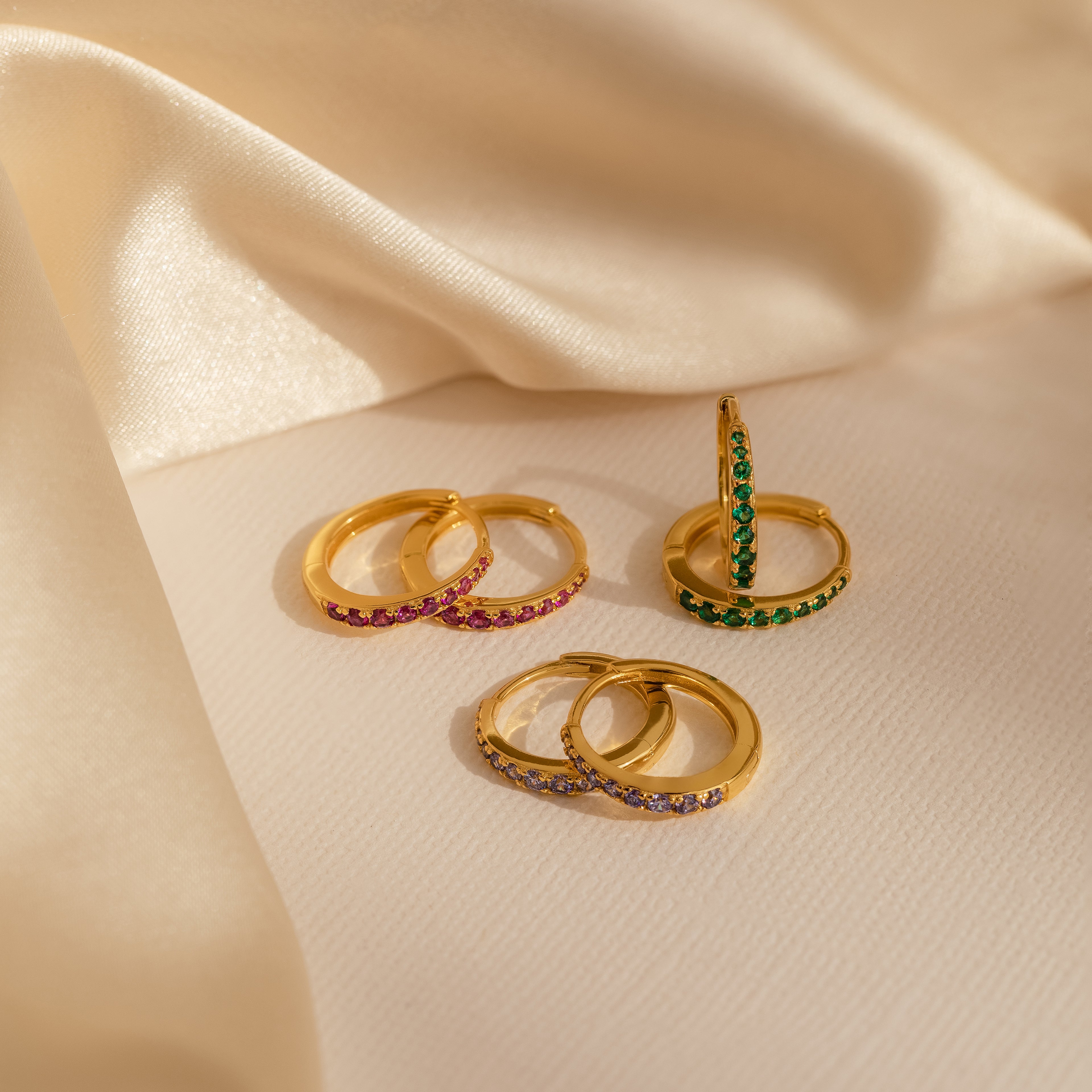 Three pairs of Vera Pave Emerald Huggies with gold hoops and green stones rest on a beige satin fabric background.