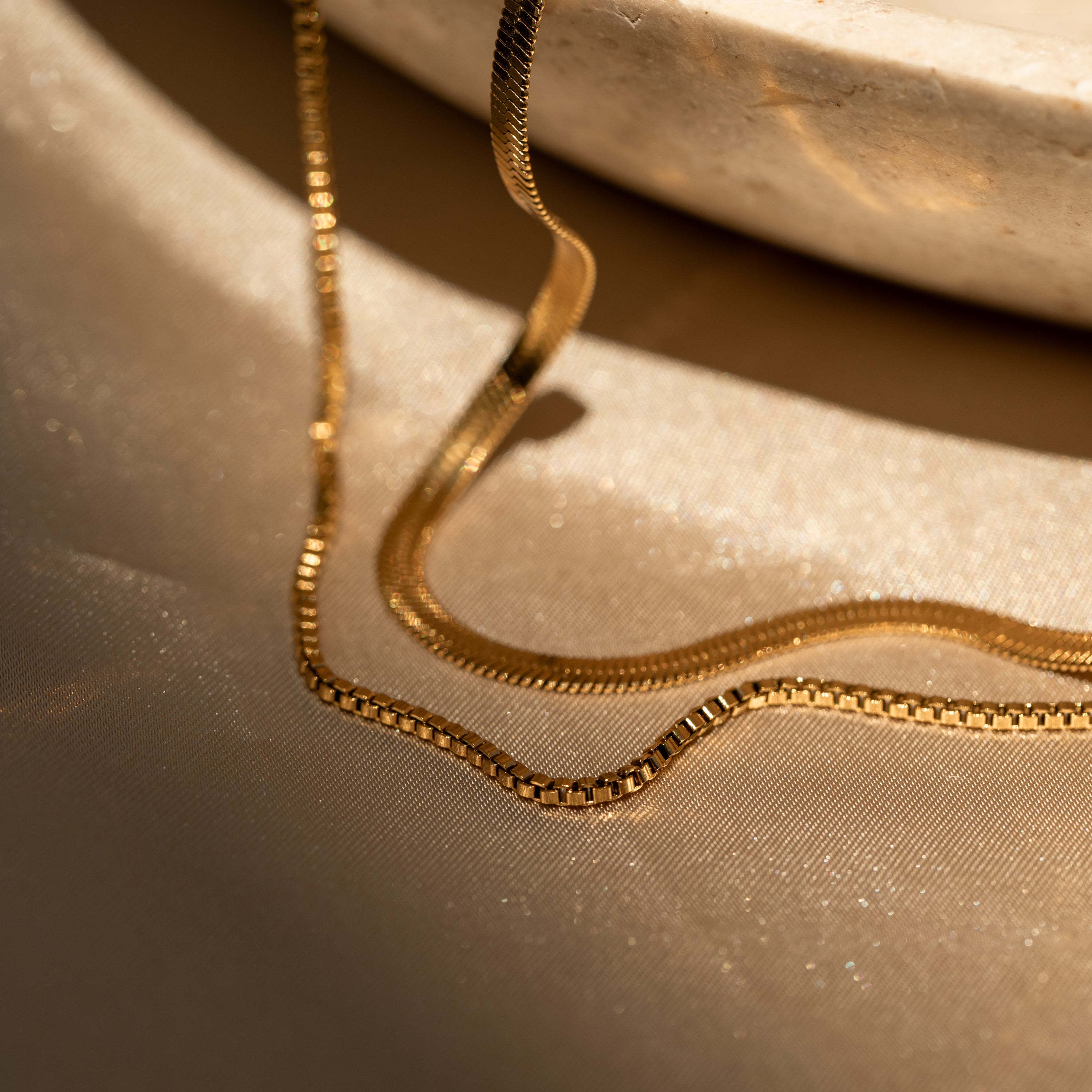 The Herringbone Duo Chain Necklace in 18K Gold rests on shimmering beige fabric beside a round marble dish.