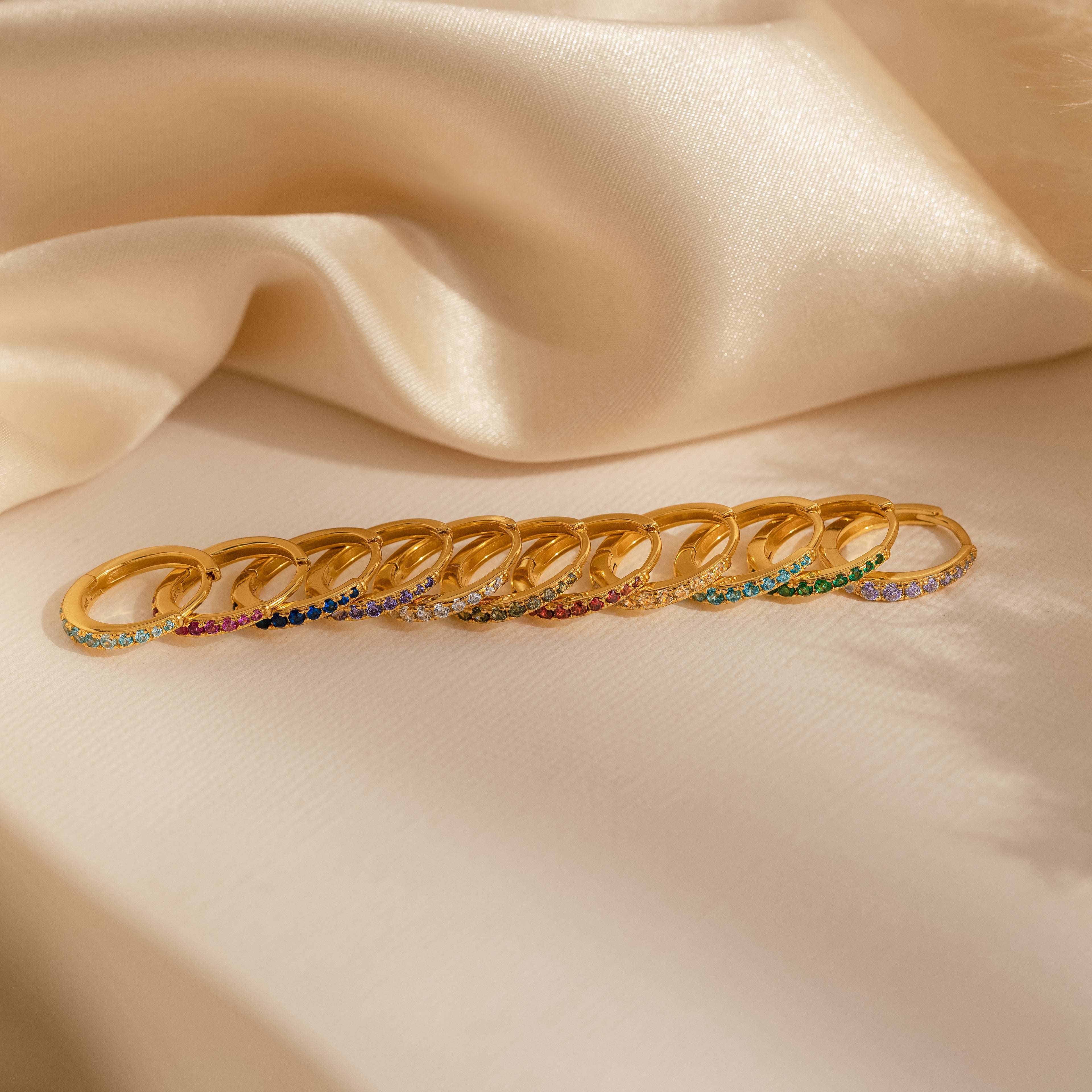 Nine Vera Pave Emerald Huggies, featuring sparkling gemstones, are arranged in a row on cream-colored satin fabric.